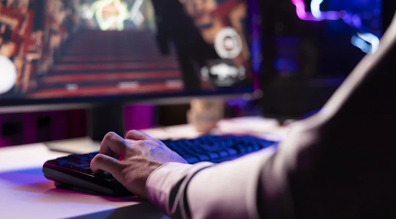 Perspetiva em primeira pessoa de um jogador a utilizar um teclado mecânico enquanto joga um jogo de PC num monitor de grandes dimensões com iluminação RGB colorida, numa configuração moderna para jogos.