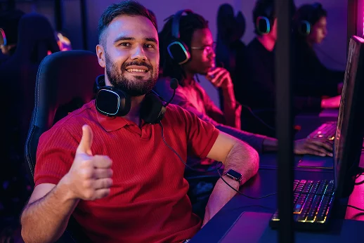 Um jogador sorridente a fazer o sinal de «polegar para cima», com auscultadores de jogos, num clube de jogos moderno, com os colegas de equipa a competir ao fundo.